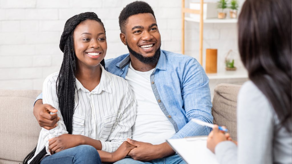 A smiling couple sits closely together on a couch, engaged in a supportive therapy session